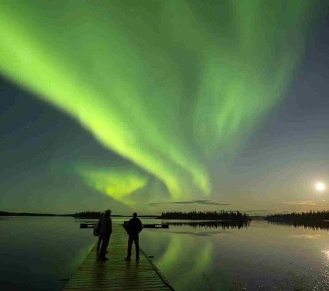 Two people standing on a dock during a full moon, watching the northern lights overhead.
